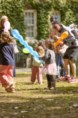 A child holds a long blue balloon in an outdoor space surrounded by other adults and children