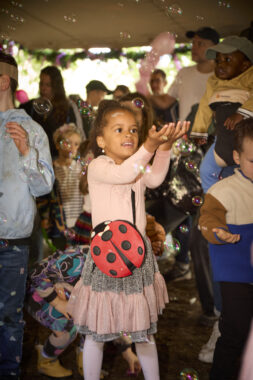 A child in a pink dress with a ladybird bag holds her hands out to catch bubbles and adults and children dance behind.