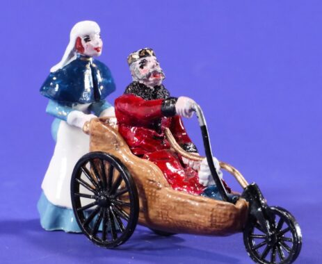 A glossy, painted figurine showing a nurse in a flowing blue cloak and white bonnet pushing a regal-looking elderly man seated in an old-fashioned wooden Bath chair. The chair features large spoked rear wheels and a smaller front wheel, with a high, curved backrest and carved brown frame. The man wears red and gold garments with a high collar and cape-like shoulders, and holds a thin cane across his lap. His expression is calm and dignified, suggesting a character of authority or nobility.