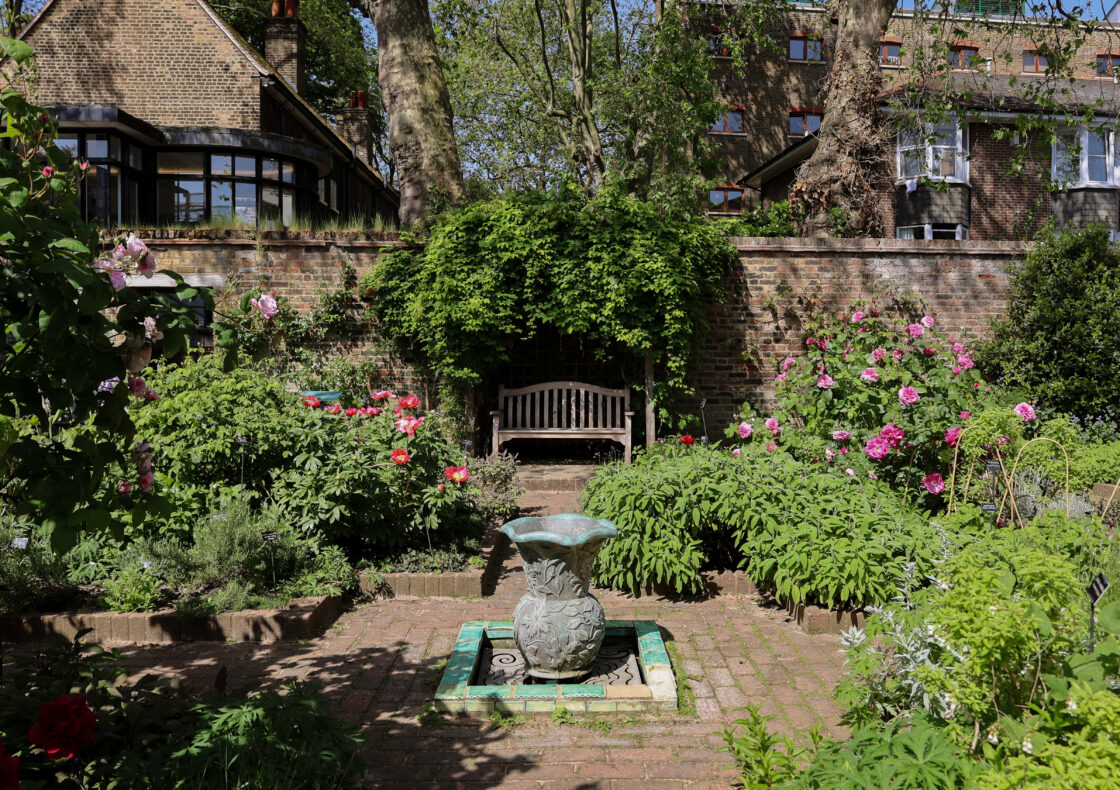 A brick garden path leads to a wooden bench beneath a leafy canopy, set against a brick wall. The garden features multiple flower beds filled with greenery and flowers. A decorative stone fountain sits at the centre of the garden.