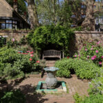 A brick garden path leads to a wooden bench beneath a leafy canopy, set against a brick wall. The garden features multiple flower beds filled with greenery and flowers. A decorative stone fountain sits at the centre of the garden.