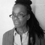 A grey-scale photo of a black woman looking to the left. She is wearing glasses, a shirt and blazer.