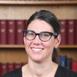 A person with long, dark hair tied up and glasses smiles at the camera in front of a book case.