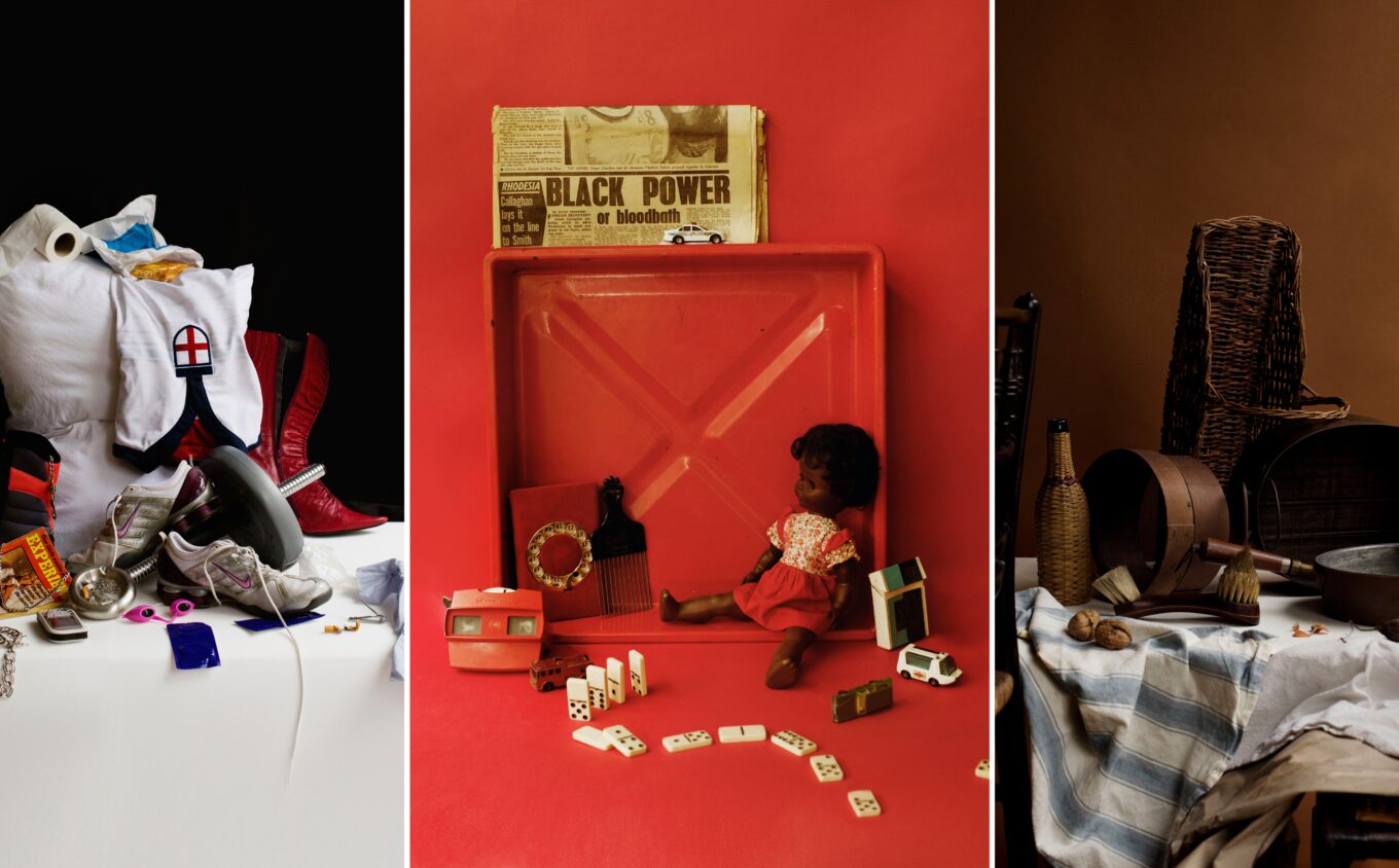 This image shows three distinct still life arrangements displayed side by side. On the left Modern items including a white pillow, red boots, england shirt, and other items tumble down. In the centre, toys, dolls, dominoes, and a newspaper reading "Black Power or bloodbath" arranged in a red display case. On the right, woven baskets, pottery, and wooden implements on cloth against a brown background.