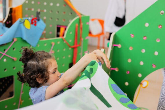 A young child clips a patterned fabric panel to a bright green pegboard playhouse frame, reaching up to secure the cloth through one of the round holes with a green clip; in the background, more fabric pieces hang from the colorful, interactive structure.