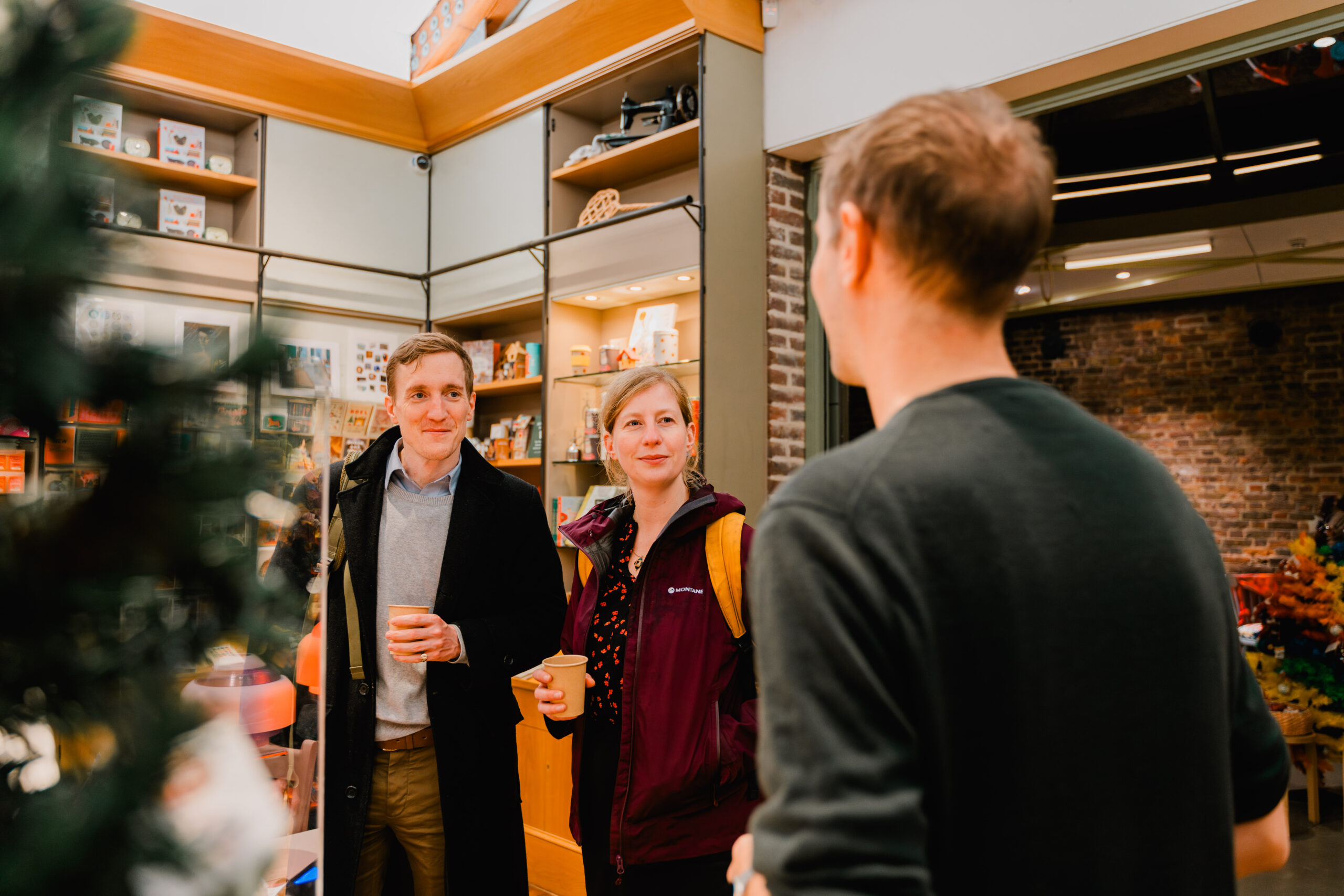 Two visitors, one in a dark overcoat and the other in a burgundy jacket with a yellow backpack, holding takeaway paper cups and smiling as they chat with a person at the museum shop where there is a Christmas tree and various shop items on the shelves.