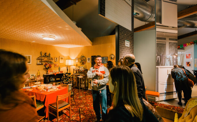 A person smiling while taking people on a tour of a 1970s room set in a gallery