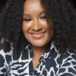 A young black woman smiles into the camera. She is wearing a black and white shirt.