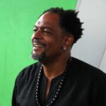 A smiling black man wearing a black shirt looks off to the left. In front of a green and white background.