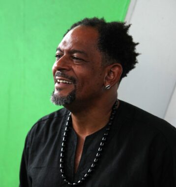 A smiling black man wearing a black shirt looks off to the left. In front of a green and white background.