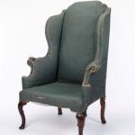 High-backed wing armchair with wooden legs on a white background. Its fully upholstered frame is covered in faded green-blue fabric, with scrolled arms and a deep seat. Some parts of the armrests are worn and fraying, revealing the underlying padding. The front legs are more slightly curved forward than the rear legs.