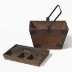 Wooden servants’ cleaning box with a folding metal handle: an inverted trapezoidal container (wider at top than base) reinforced with black metal corner straps, accompanied by a shallow tray with six compartments and a central cut-out handle.