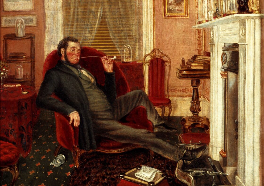 Victorian parlour: a man in a dark suit and frock coat reclines in a deep red velvet armchair, legs outstretched with one foot on the brass fireplace fender, drawing on a long, slender pipe. There are lace curtains by the window behind him; a small table piled with books on the right; framed artworks hang on the walls; a footstool and scattered playing cards lie on the patterned carpet.