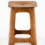 Warm honey-toned bent birch plywood stool in a white background: square seat with rounded corners and a shallow, bowl-like depression at its centre, held in place by a central bolt. Its trapezoidal base flares outward into four rounded legs, each pierced with a large rectangular.
