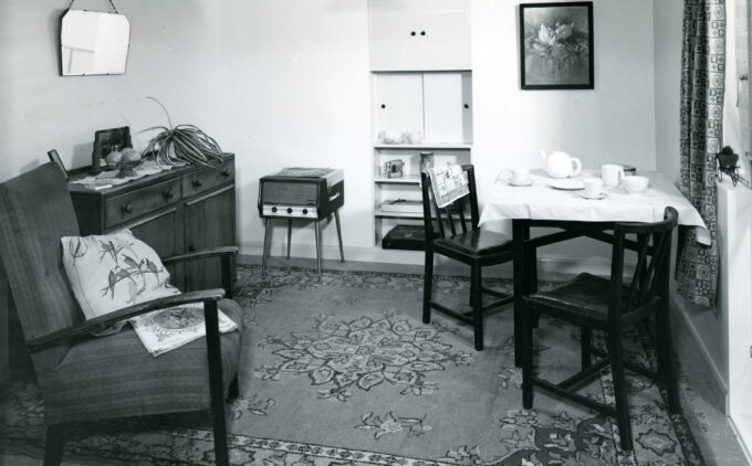 A black and white image of a compact 1950s council flat living room: a patterned rug covers the floor, an upholstered armchair with cushions sits beside a low sideboard, and a small square table set for tea—with cups, saucers, and a teapot— with two wooden chairs. Built-in shelving and a storage cupboard on the back wall holds various small objects.