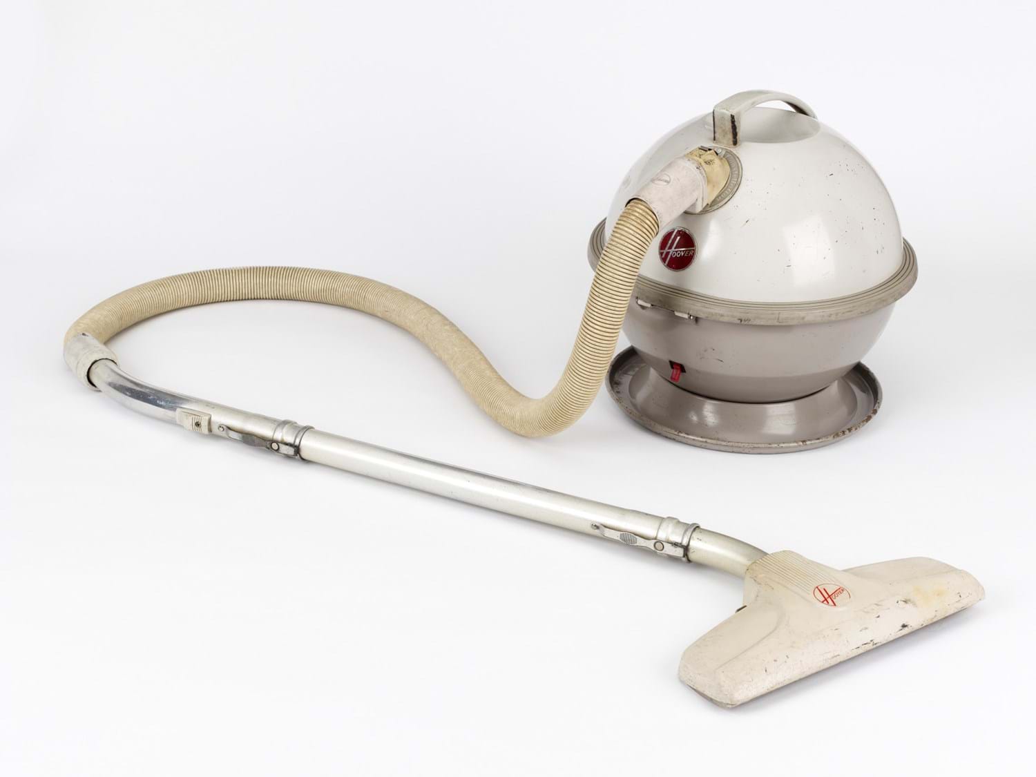 White-and-beige spherical Hoover Constellation vacuum cleaner on a white background: its ball-shaped body with a carry handle and red Hoover logo feeds a flexible beige hose and straight silver tube, ending in a white plastic floor nozzle.