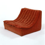 Low-slung scoop chair on a white background, upholstered in rust-coloured cotton corduroy with vertical channel quilting.