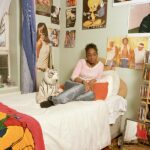 A teenage girl sits on the bed with her eyes closed in a brightly lit bedroom. The bed has a white coverlet and a red Pooh-themed throw blanket; beside her is a white tiger soft toy behind her, pale green walls are plastered with posters and photographs, and a wooden shelf holds folded clothes, CDs and a red alarm clock. A lace-curtained window with a teddy bear on its sill fills the left side of the frame.