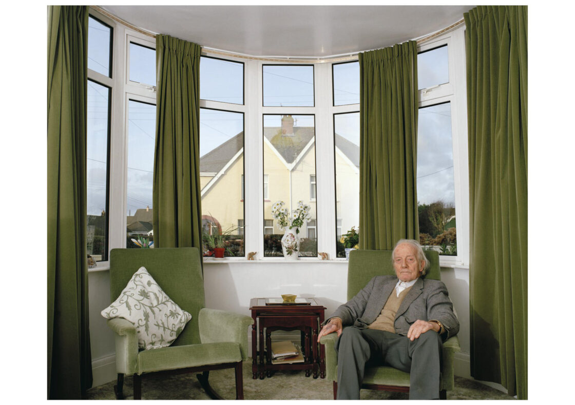An elderly man sits in the right of two matching sage green armchairs inside a curved bay window alcove with matching green floor length curtains. He rests his hands on the arms while gazing blankly into the air. Between the chairs stands a small, dark wooden nest of tables.