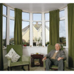 An elderly man sits in the right of two matching sage green armchairs inside a curved bay window alcove with matching green floor length curtains. He rests his hands on the arms while gazing blankly into the air. Between the chairs stands a small, dark wooden nest of tables.