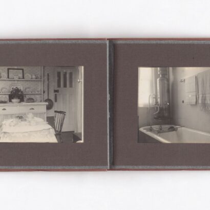 Open vintage soft brown cloth photo album showing sepia toned photos: a tea-set table before a plate-laden sideboard on the left, and a bathtub with two towels on rails and a window on the right.