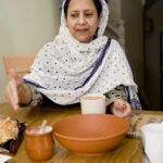 A middle-aged woman wearing a white headscarf dotted with blue sits at a wooden table, extending her hand toward a large terracotta bowl; on the table before her are a small clay pot with a spoon, a white mug, and a plate of snacks.