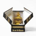 Black presentation box on a white background, its flaps open to reveal a small ornate gold perfume bottle. On the box is gold script and patterns including "Attar Al Kaaba"