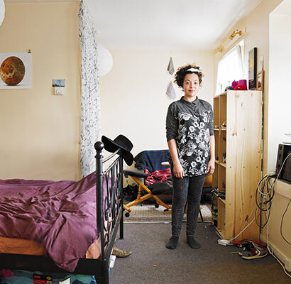 Compact cream‐walled room with a black metal bed with a rumpled plum-purple duvet under a white paper ceiling light shade on the left; In the centre, a young person is standing. On the right, a tall pine shelving unit and a window-side area hold a black printer, other electronics, and coiled cables.