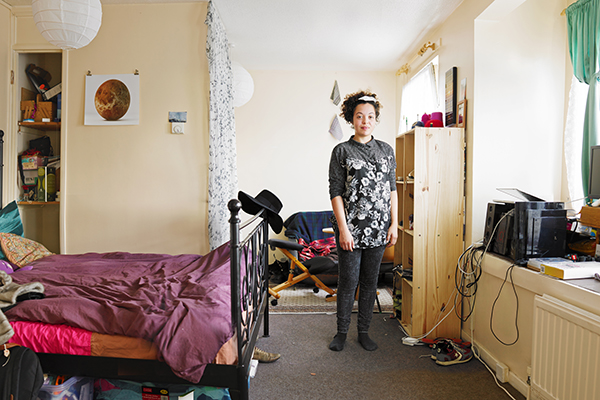 Compact cream‐walled room with a black metal bed with a rumpled plum-purple duvet under a white paper ceiling light shade on the left; In the centre, a young person is standing. On the right, a tall pine shelving unit and a window-side area hold a black printer, other electronics, and coiled cables.
