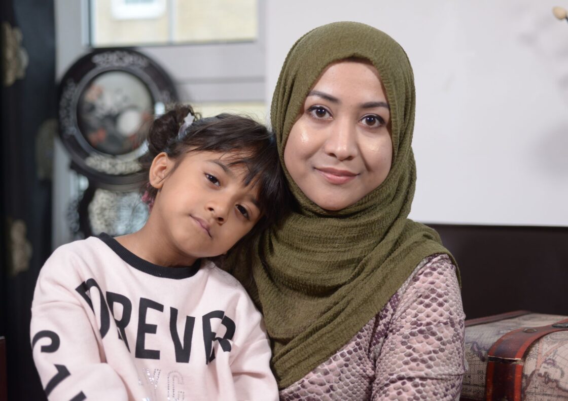 A close-up of a mother with her young daughter leaning on her shoulder looking straight into the camera; behind them, glimpses of their domestic space.
