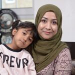 A close-up of a mother with her young daughter leaning on her shoulder looking straight into the camera; behind them, glimpses of their domestic space.