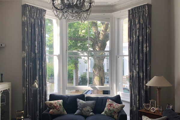 Bay window with blue patterned curtains, navy sofa with floral cushions, and chandelier.