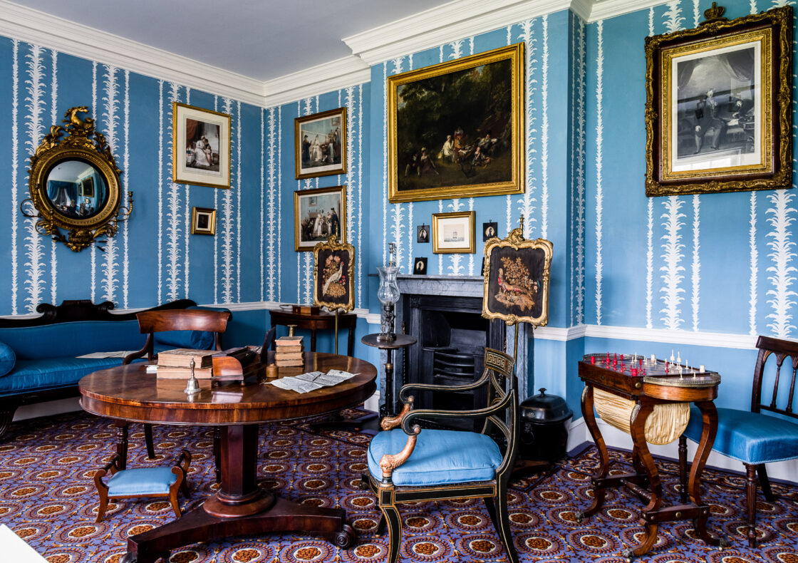 Room with blue decorative wallpaper, multiple paintings and a round mirror in gilt frames, a dark marble fireplace behind a round table atop sit books, a bell and a letter in the process of being written. There are dark wood chairs with blue cushioning, and decorative fire screens depicting pheasants.