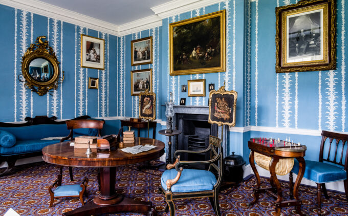 Room with blue decorative wallpaper, multiple paintings and a round mirror in gilt frames, a dark marble fireplace behind a round table atop sit books, a bell and a letter in the process of being written. There are dark wood chairs with blue cushioning, and decorative fire screens depicting pheasants.