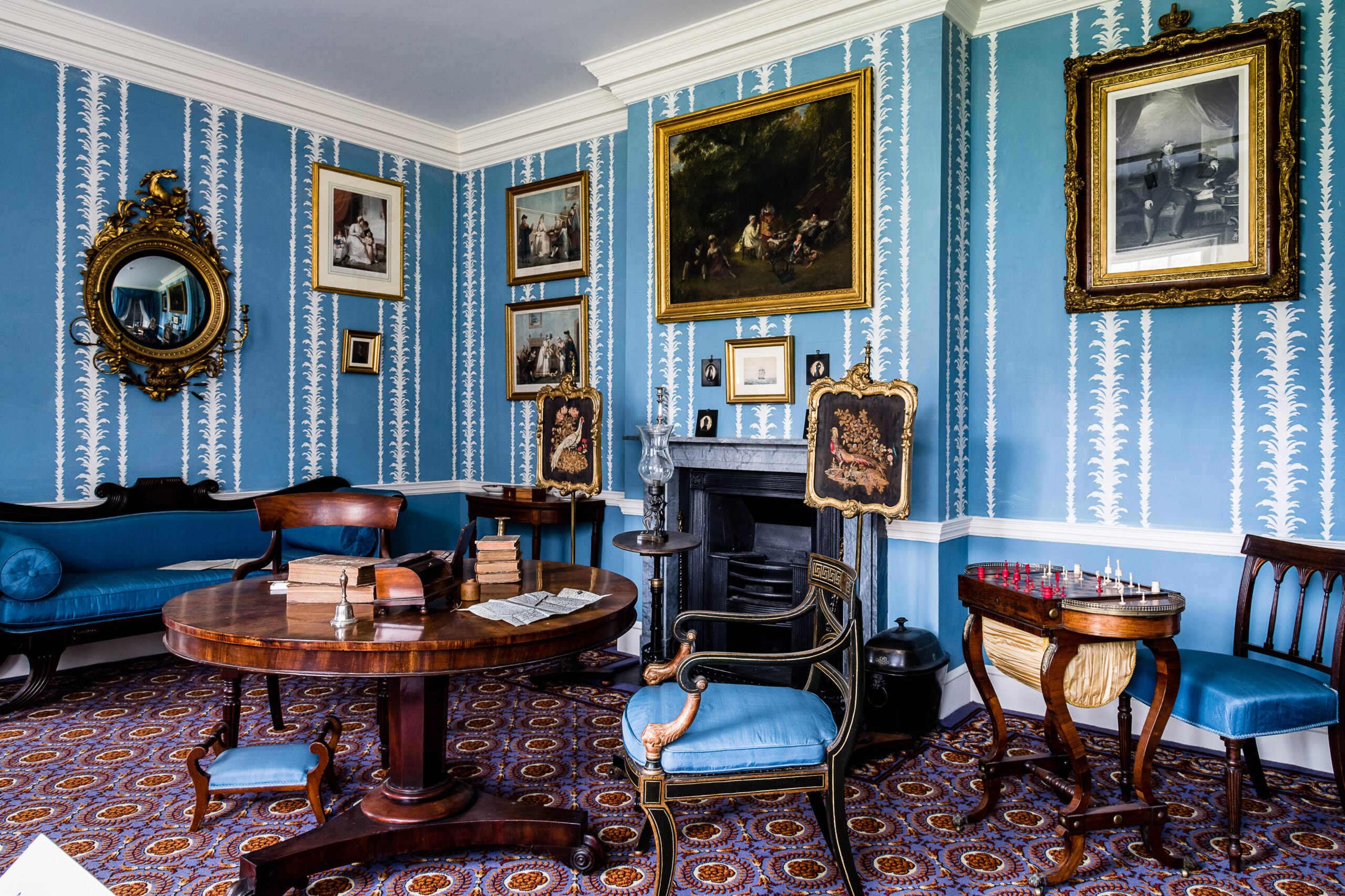 Room with blue decorative wallpaper, multiple paintings and a round mirror in gilt frames, a dark marble fireplace behind a round table atop sit books, a bell and a letter in the process of being written. There are dark wood chairs with blue cushioning, and decorative fire screens depicting pheasants.