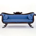 Dark wood settee with blue upholstery on tapered legs with curved arms and decorative carved details.