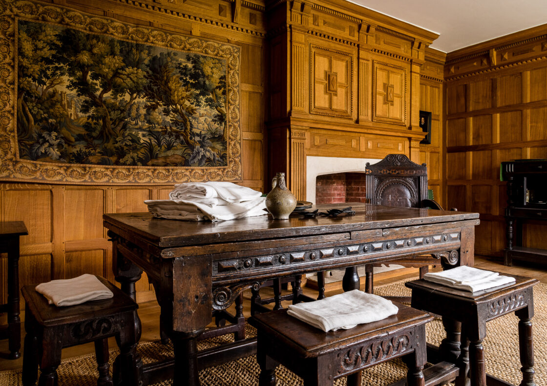 A wood-paneled room with ornate tapestry, wooden table and stools, fireplace, and woven rug.
