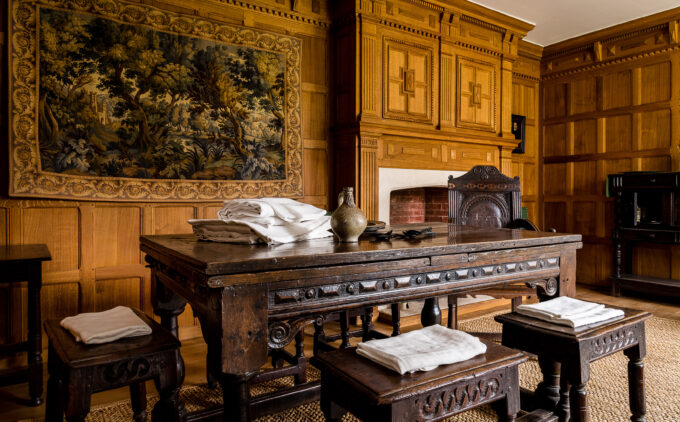 A wood-paneled room with ornate tapestry, wooden table and stools, fireplace, and woven rug.