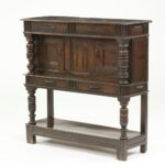 Oak court cupboard with carved panels, turned columns, and multiple drawers on tall legs with lower shelf.