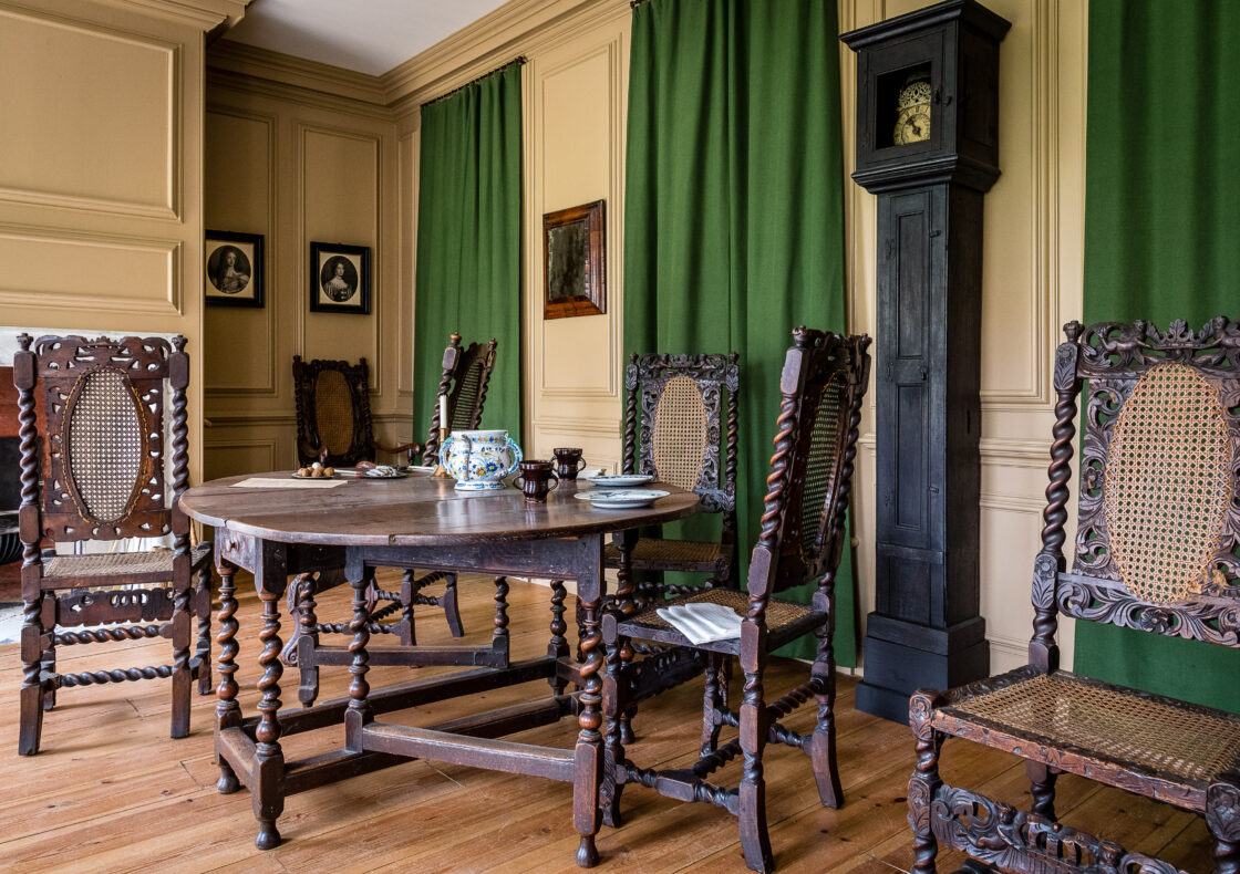 Room with carved wooden chairs, round table, tall grandfather clock, green curtains, and cream paneled walls.