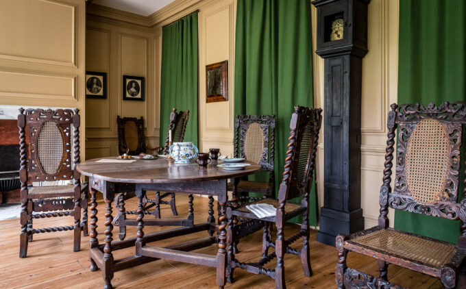 Room with carved wooden chairs, round table, tall grandfather clock, green curtains, and cream paneled walls.