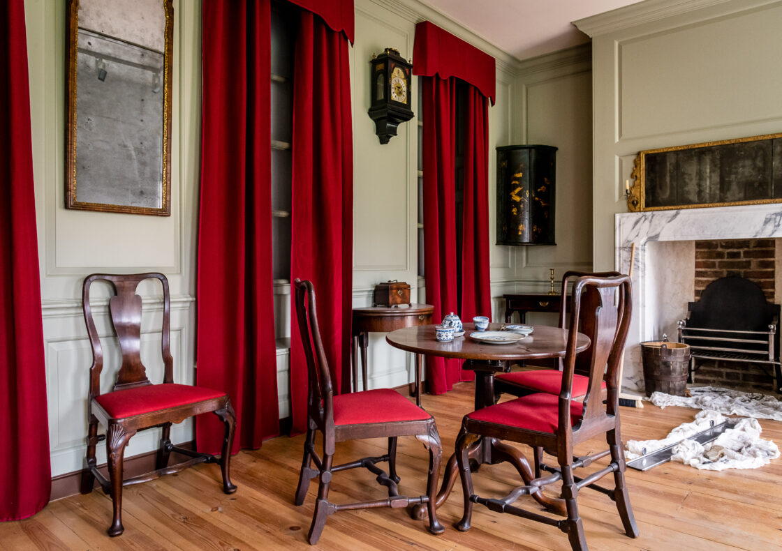 A parlour with wooden chairs with red cushions around a small round table. Rich red curtains are against a pale green wall. A fireplace with marble surround is visible on the right, other details include a wall-mounted clock and mirrors.