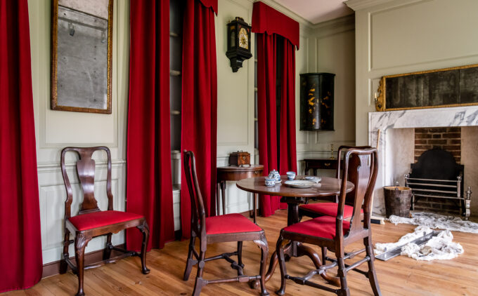 A parlour with wooden chairs with red cushions around a small round table. Rich red curtains are against a pale green wall. A fireplace with marble surround is visible on the right, other details include a wall-mounted clock and mirrors.