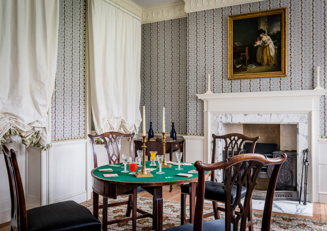 Room with green-topped card table, chairs, marble fireplace, patterned wallpaper, white curtains, and gilt-framed portrait above mantel.
