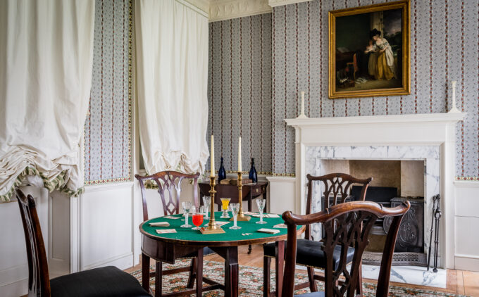 Room with green-topped card table, chairs, marble fireplace, patterned wallpaper, white curtains, and gilt-framed portrait above mantel.