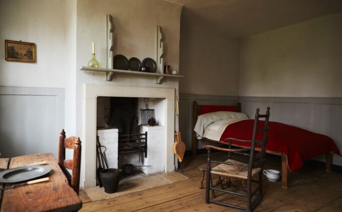An almshouse room with wide floorboards and simple furnishings: on the left is a wooden table set with a plate and knife; in the centre is a white‐painted fireplace with iron fire tools and a mantel shelf displaying three plates a single candle in a glass bottle; and on the right is plain single bed draped in a red blanket sits beside a ladder‐back chair.