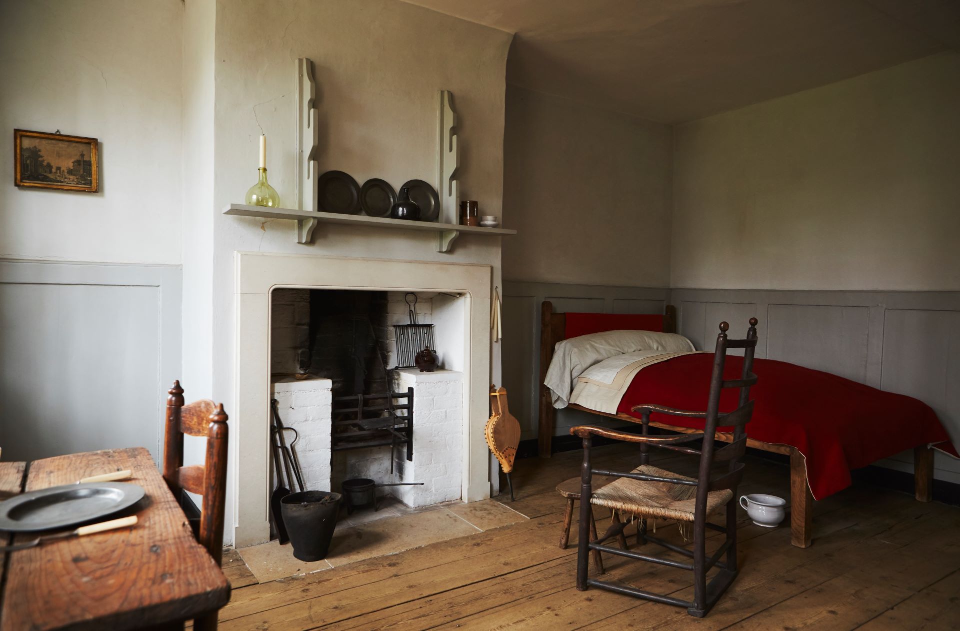 An almshouse room with wide floorboards and simple furnishings: on the left is a wooden table set with a plate and knife; in the centre is a white‐painted fireplace with iron fire tools and a mantel shelf displaying three plates a single candle in a glass bottle; and on the right is plain single bed draped in a red blanket sits beside a ladder‐back chair.