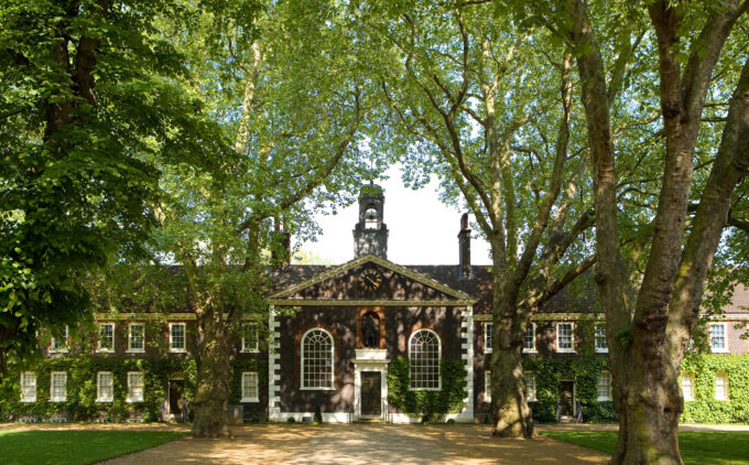 A symmetrical red-brick almshouse framed by the canopy of mature plane trees. At its centre is a pedimented entrance with a black door flanked by tall arched windows; above the doorway, a niche houses a statue of Robert Geffrye, and a clock adorns the triangular pediment above that. Ivy climbs the façades of the both wings of the building, while dappled sunlight patterns the gravel path and manicured lawn in the foreground.