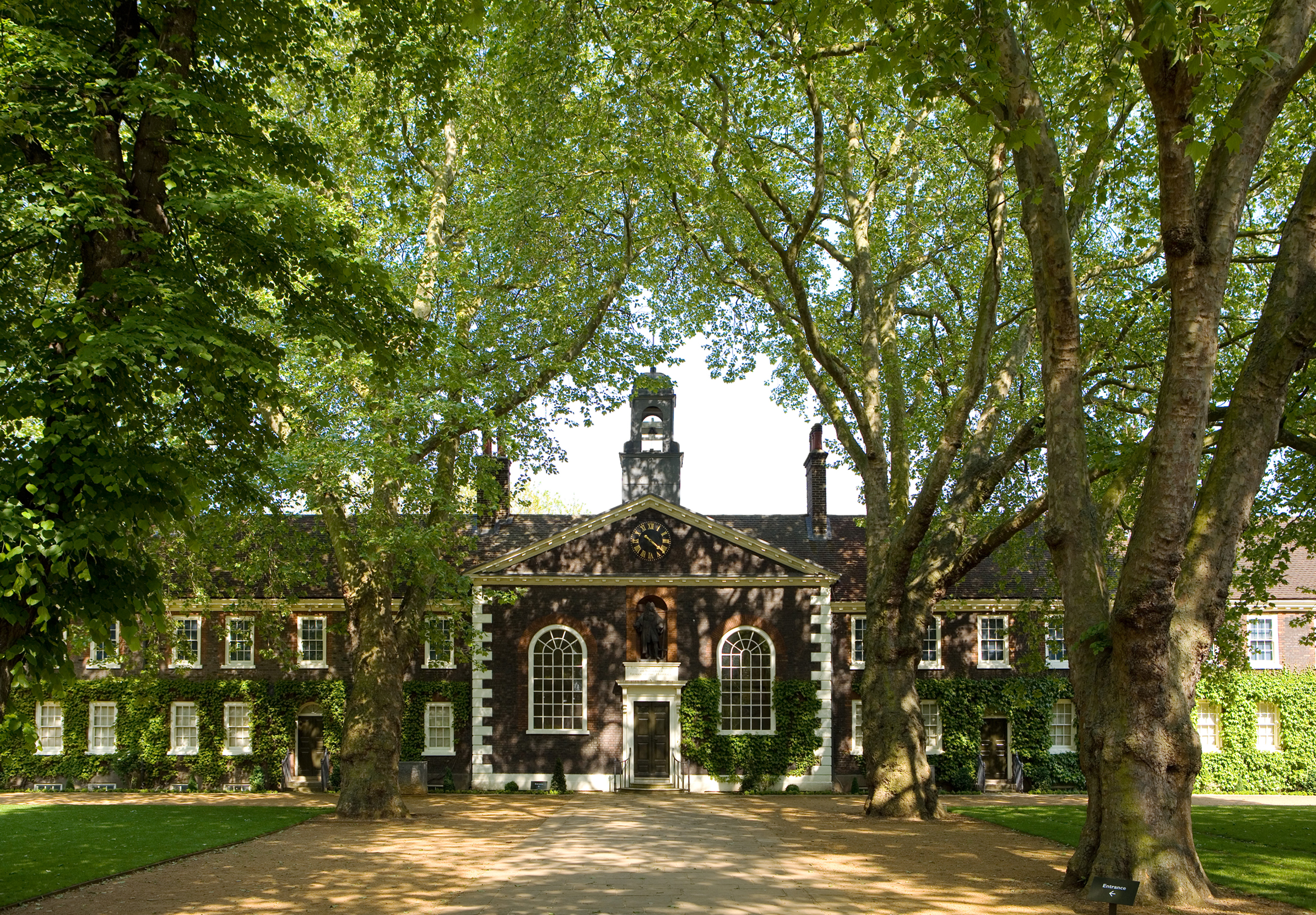 A symmetrical red-brick almshouse framed by the canopy of mature plane trees. At its centre is a pedimented entrance with a black door flanked by tall arched windows; above the doorway, a niche houses a statue of Robert Geffrye, and a clock adorns the triangular pediment above that. Ivy climbs the façades of the both wings of the building, while dappled sunlight patterns the gravel path and manicured lawn in the foreground.