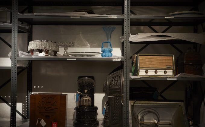 A dimly lit steel shelving unit lined with white tissue paper and labelled with small white stickers. The shelves hold stored objects including a shallow glass platter, plates, a blue glass basket, a tall flared blue glass vase, a pale yellow vintage radio, a wooden radio cabinet, and more.